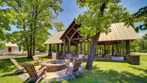 Um pavilhão ao ar livre com cadeiras Adirondack, uma lareira e uma área coberta para sentar, tudo cercado por árvores.