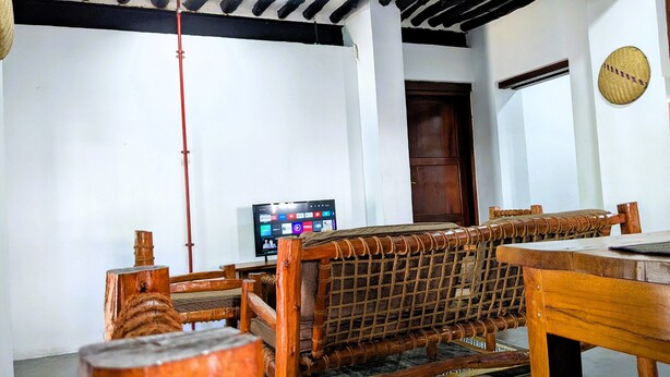 A traditional wooden room with a woven bench, a television displaying streaming services, and a wooden door. - The Hub Train House Amazing Gents Code (Zanzibar Town)