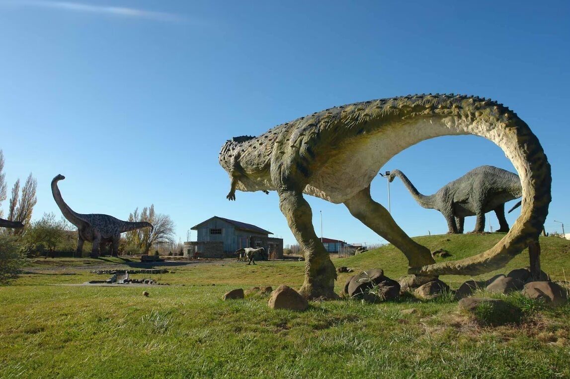 A dinosaur exhibit with large models of sauropods, a grassy field, and a building in the background.