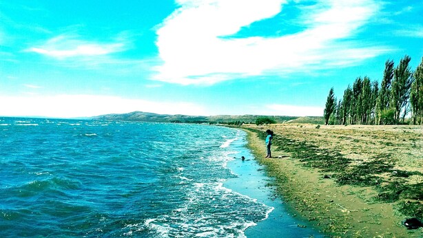 Un paisaje costero con playa, olas del océano y una persona de pie junto al agua.