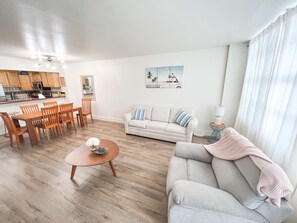 A living room with a sofa, coffee table, dining table, and chairs. There is a kitchen area with cabinets and a microwave. - Elegant 1BR Oceanfront Retreat Sleeps 6 (Hollywood)