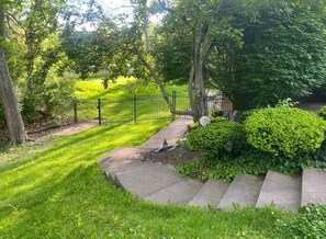 A residential outdoor area with a paved pathway, a metal fence, and lush greenery. - Broad Ripple Retreat | Private Gym & Yard (Indianapolis)