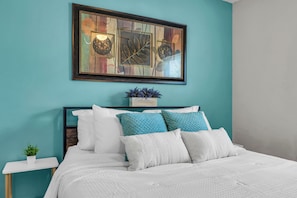 A neatly made bed with white bedding and teal pillows, a small potted plant on a bedside table, and a framed artwork above the bed. - Hidden Gem!  3BR Family home w/ Fire Pit & Games (St. Augustine)