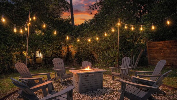 Un espacio al aire libre con sillas Adirondack y un hogar para fogata, rodeado de árboles y luces colgantes.