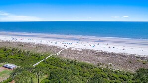 En vy över en strand från ovan, med många parasoller och människor, omgiven av grönska och en klar himmel.