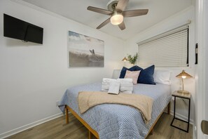 Ein Schlafzimmer mit einem Bett, einem Deckenventilator, einem Fernseher, einem Fenster mit Jalousien und einem Nachttisch mit Lampe.