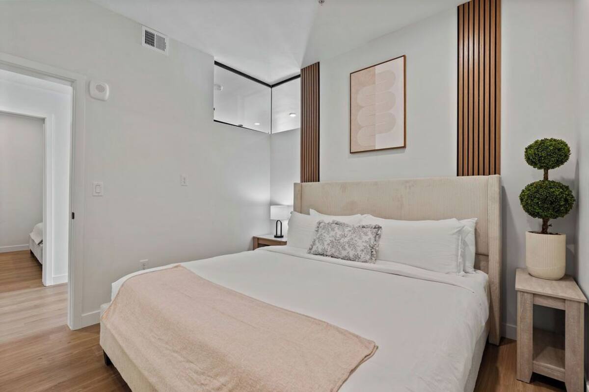 A bedroom with a large bed, a bedside table, a nightstand, a wall-mounted mirror, and a potted plant.
