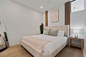 Quarto moderno com uma cama grande, mesa de cabeceira com abajur, obra de arte na parede e um vaso com planta.