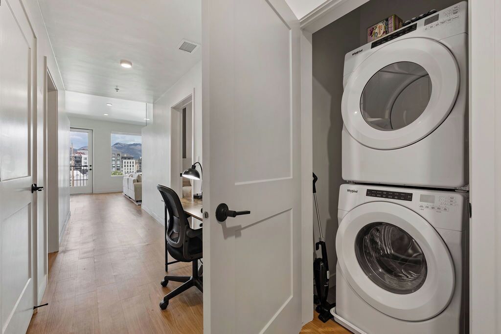 A laundry room with two stacked washing machines, a desk with a chair, and a view of a cityscape through an open door.