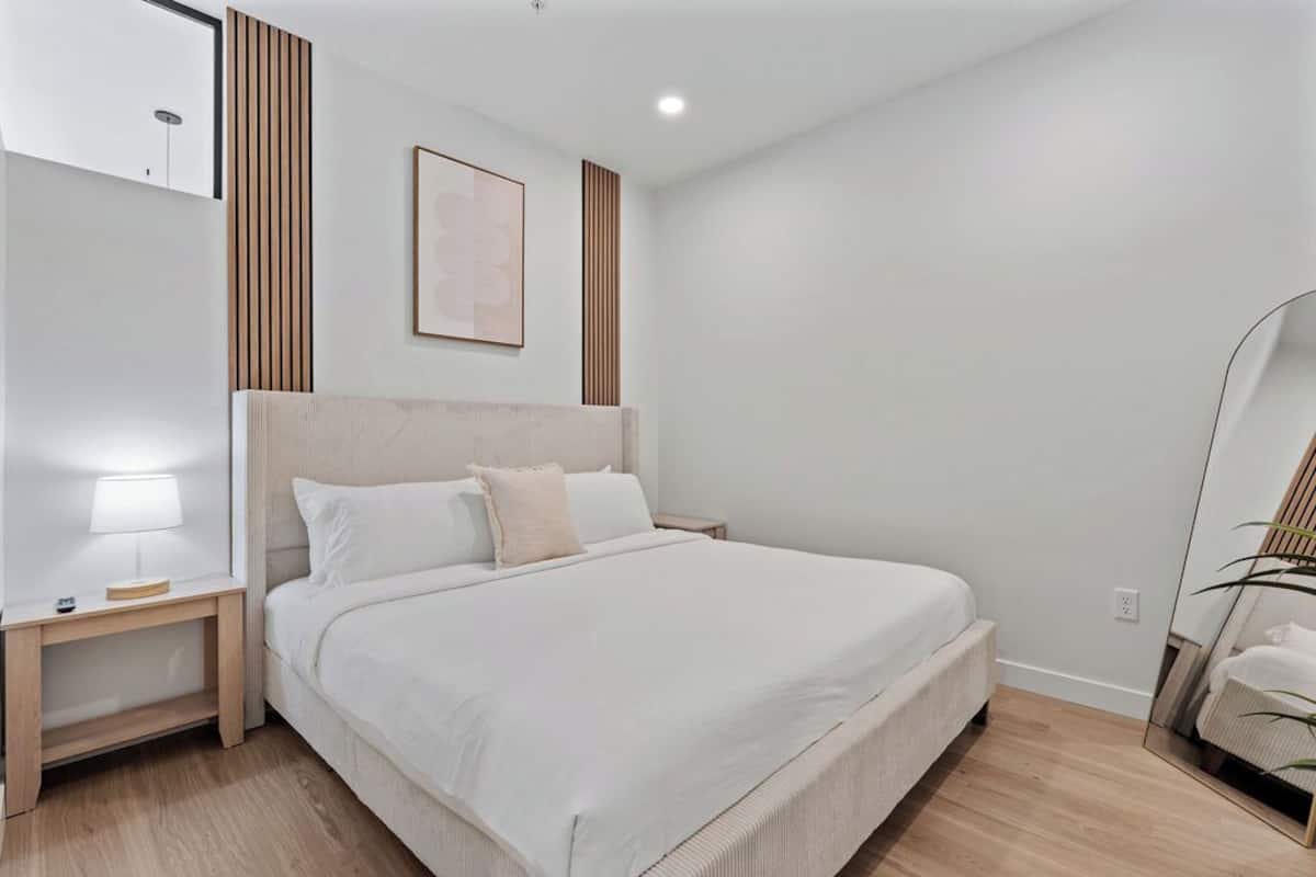 A bedroom with a large bed, a bedside table, a nightstand, a wall-mounted light, and a framed picture.