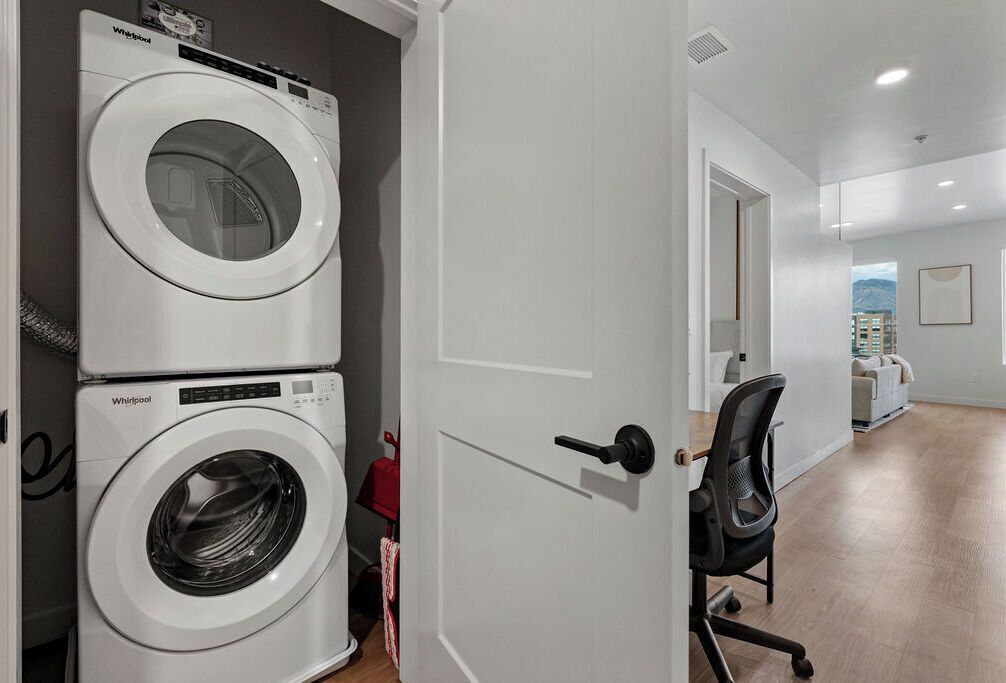 A laundry room with two stacked Whirlpool washers, a door leading to a hallway, and a chair in the room.