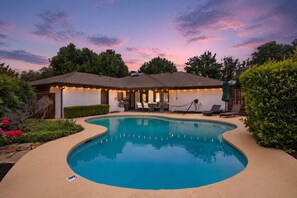 Ein Pool mit gewölbter Kante, umgeben von einer gepflasterten Liegefläche. Liegestühle und ein Grillbereich sind vorhanden.