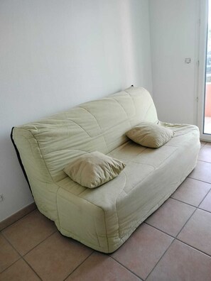 A light-colored sofa with two cushions in a room with tiled flooring and a plain wall. - Alexandre's southern apartment with covered terrace, swimming pool and private garage (Saint-Raphaël)