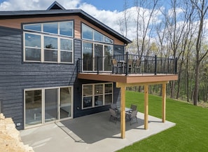 A modern two-story house with a balcony, large windows, and a patio with outdoor furniture.