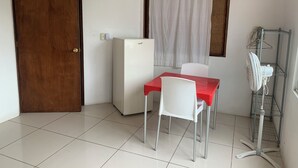 A small room with a wooden door, a white refrigerator, a red table with a white chair, a white curtain, and a metal rack with a fan. - BRISAMAR GUESTHOUSE (Puerto Escondido)