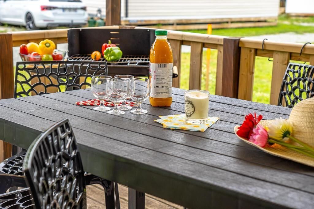 Una terraza de madera con una mesa dispuesta para una comida, que incluye una botella de jugo, vasos y un sombrero adornado con flores.