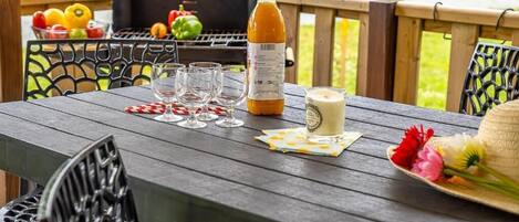 Una terraza de madera con una mesa dispuesta para una comida, que incluye una botella de jugo, vasos y un sombrero adornado con flores.