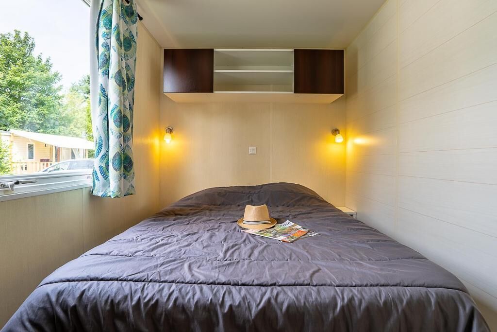 Una cama individual con un edredón oscuro, un sombrero y una revista encima. Hay dos lámparas de pared a cada lado de la cama.