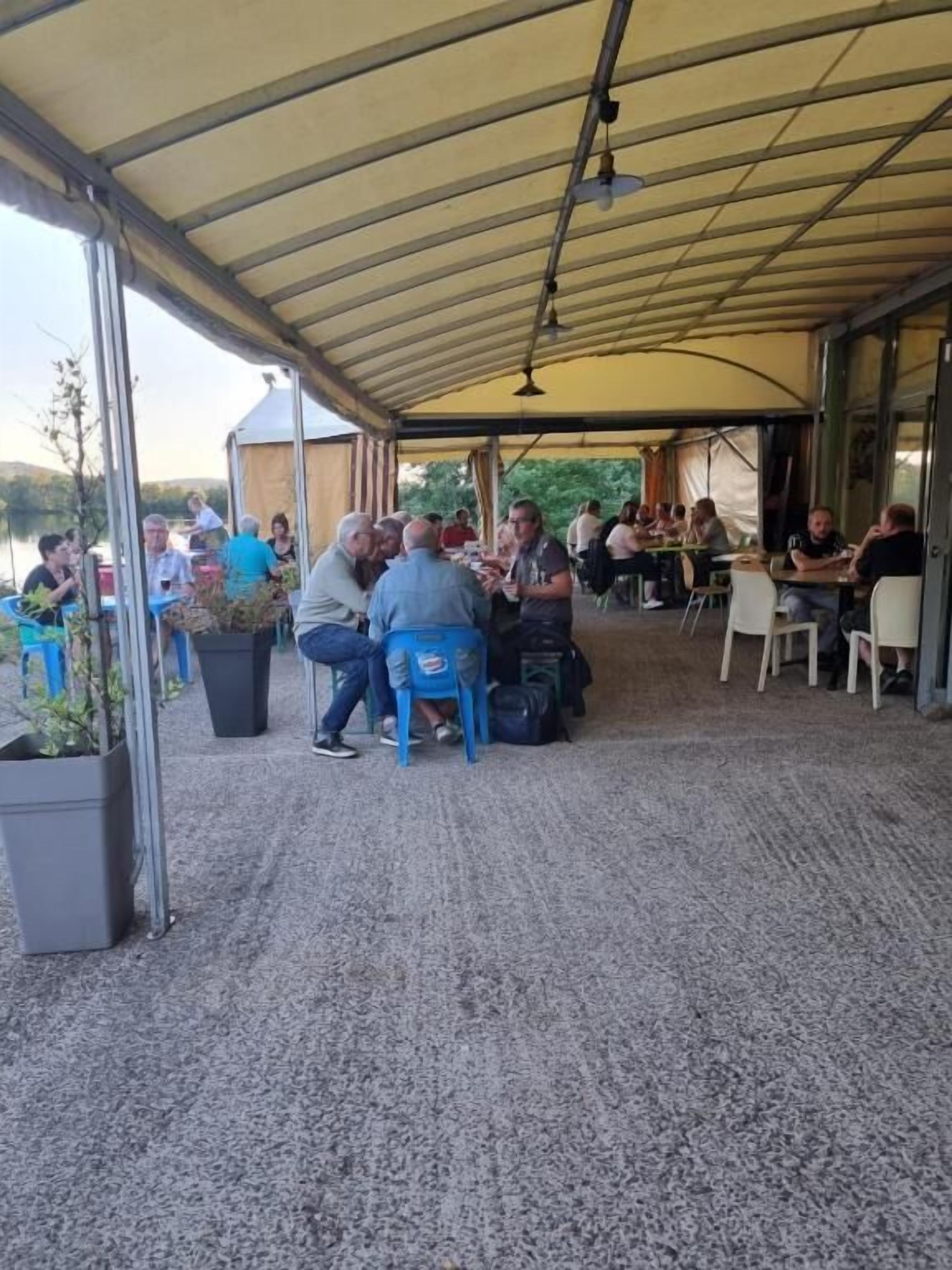 Un espace extérieur aménagé avec des tables et des chaises sous une tonnelle, entouré de plantes en pot et offrant une vue sur le lac.