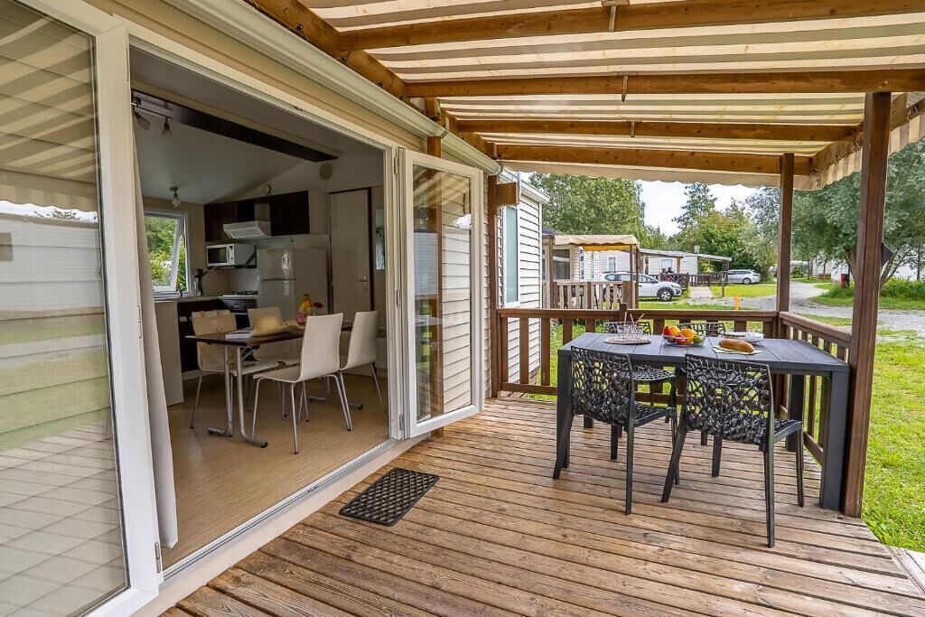 Een overdekt terras met een tafel en stoelen, een schuifdeur naar een keuken, en uitzicht op het omliggende landschap.