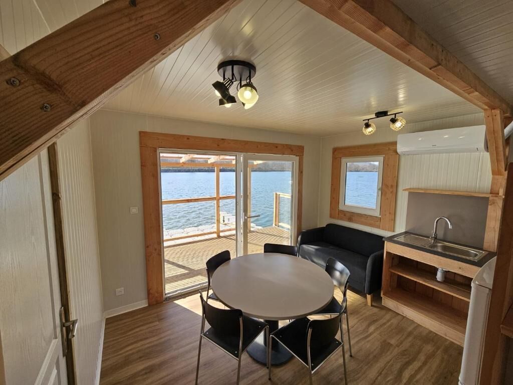 Une pièce avec un plafond en bois, une table ronde entourée de chaises, un canapé noir, un coin cuisine avec un évier et de grandes fenêtres donnant sur l’eau.