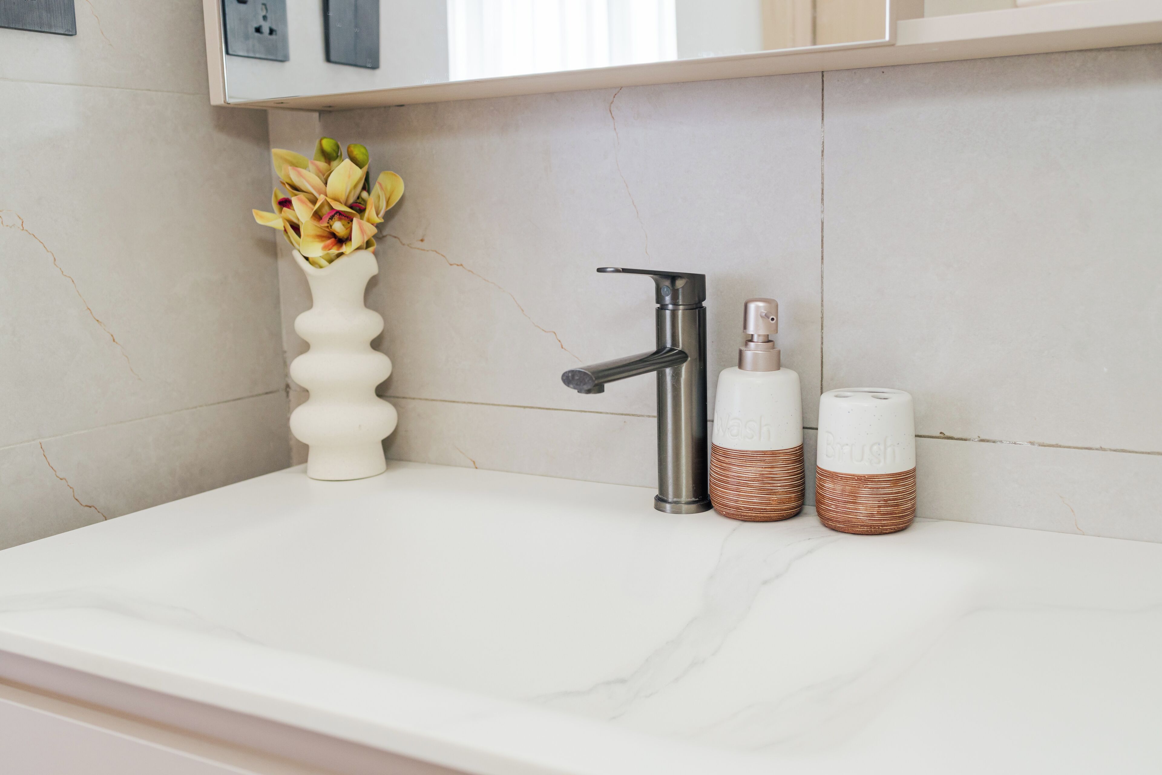 Une salle de bain avec un lavabo en marbre blanc, un robinet moderne, un vase avec des fleurs et deux flacons de produits de toilette.