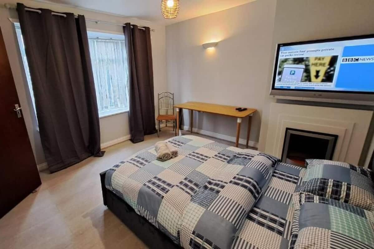 A bedroom with a bed, a desk, a chair, a television, and a fireplace.