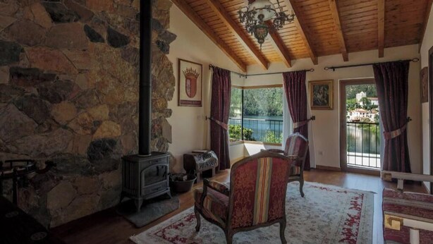 Una habitación con paredes de piedra, vigas de madera, chimenea, una silla y vista a un lago.