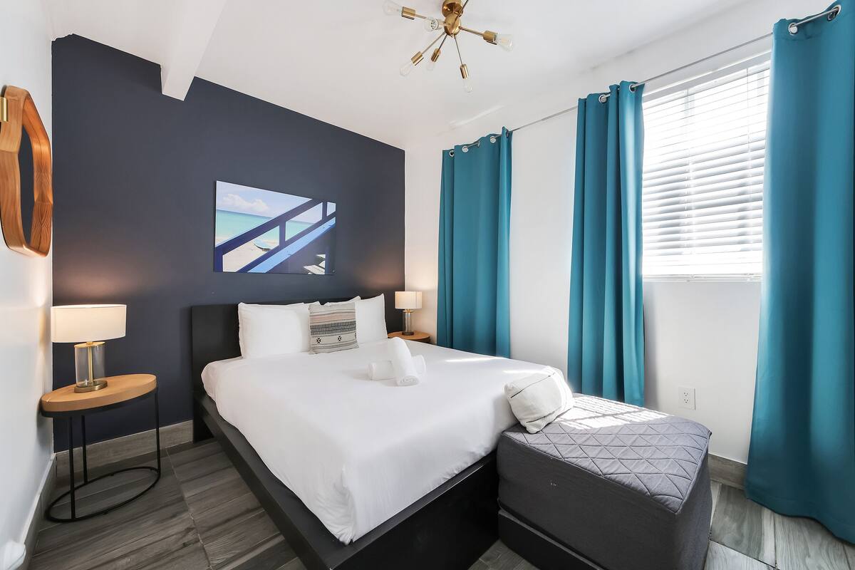 A modern hotel room with a bed, bedside tables, a nightstand, a bench, a ceiling light fixture, and a window with curtains.