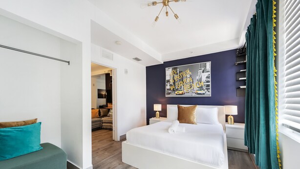 A modern hotel room with a large bed, a seating area, a painting on the wall, and a window with curtains. - Drexel Apts by At Mine Hospitality (Miami Beach)