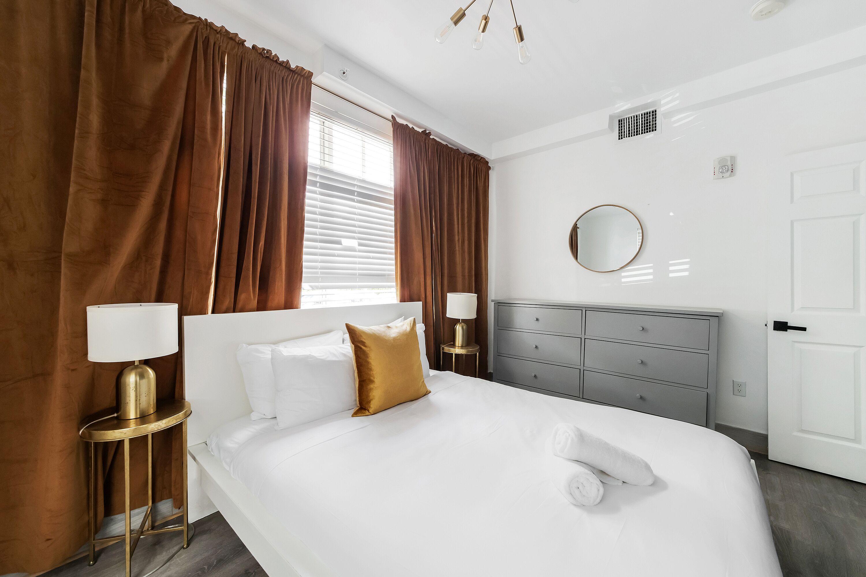 Una habitación de hotel con una cama grande, dos mesitas de noche con lámparas, un espejo redondo y una puerta.