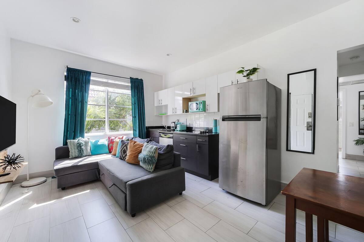 A modern living room with a sofa, a dining table, a kitchen area with cabinets and appliances, and a large window with curtains.