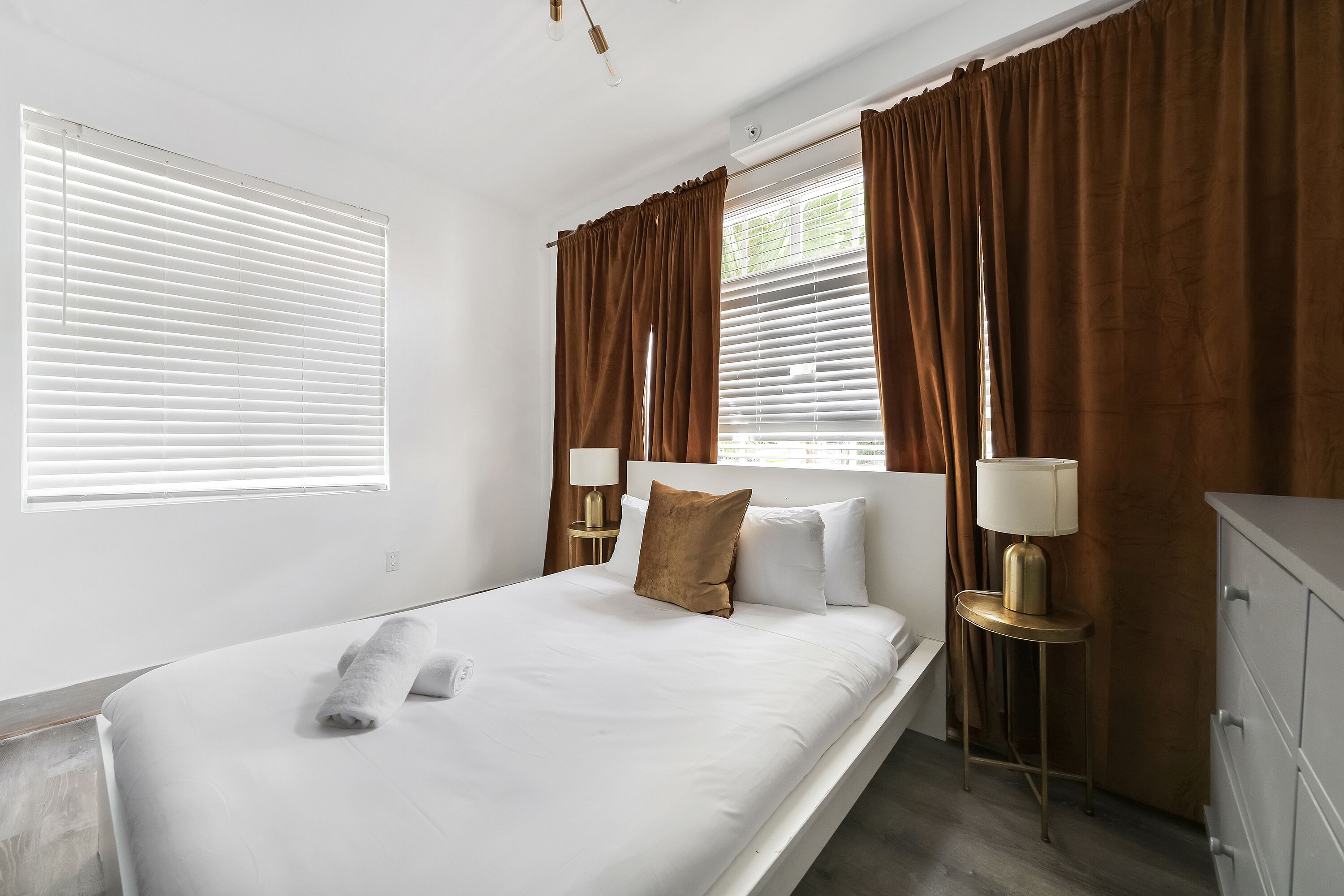 A neatly made bed with white linens and a brown pillow, a bedside lamp, and a dresser.