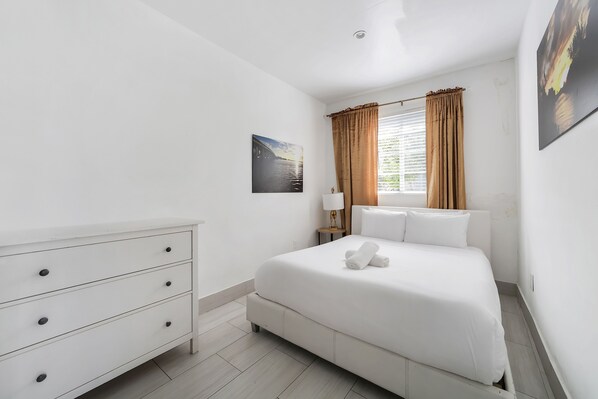 Un dormitorio con cama, cómoda blanca, mesita de noche, lámpara y ventana con cortinas.