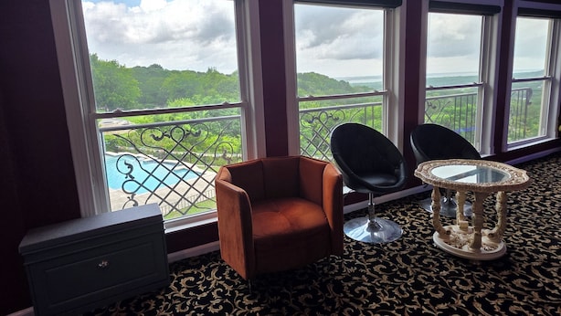 Une pièce avec vue sur une piscine et sur la verdure, un fauteuil marron, deux chaises noires, une petite table et un tapis à motifs sombres.
