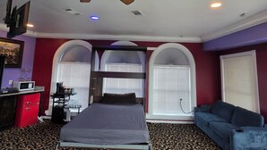 Une chambre à coucher comprenant un lit, un ventilateur de plafond, un téléviseur, un bureau avec un ordinateur et une fenêtre avec des stores.