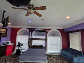 A bedroom with a bed, a ceiling fan, a TV, a desk with a computer, and a window with blinds.