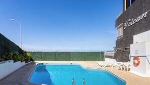 Ein Schwimmbecken mit Rettungsboje, ein weißes Gebäude mit dem Schriftzug 'Volcan' und ein Granhügel.