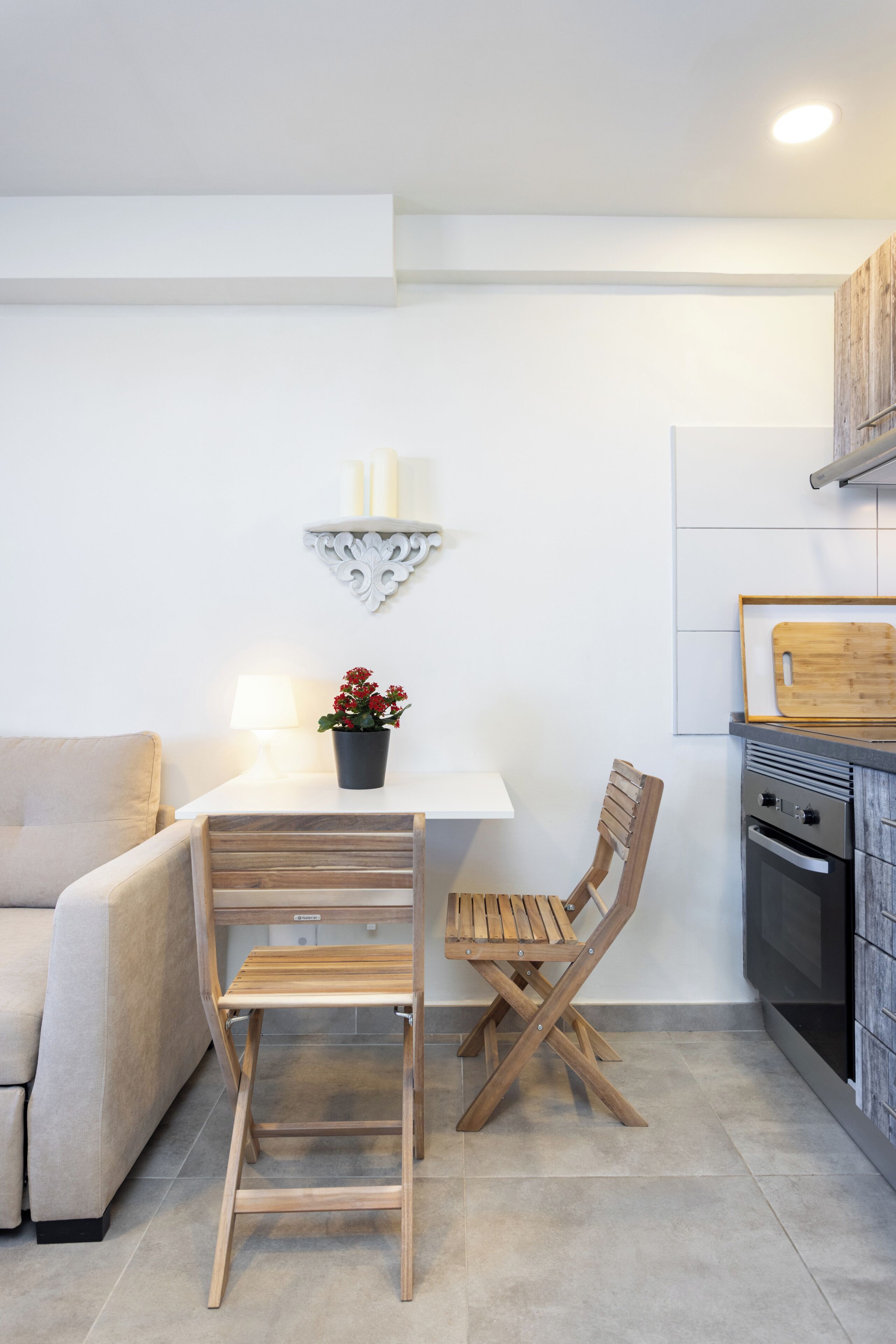 A modern kitchen with a white countertop, wooden chairs, a small table with a potted plant, and a wall-mounted shelf with candles.