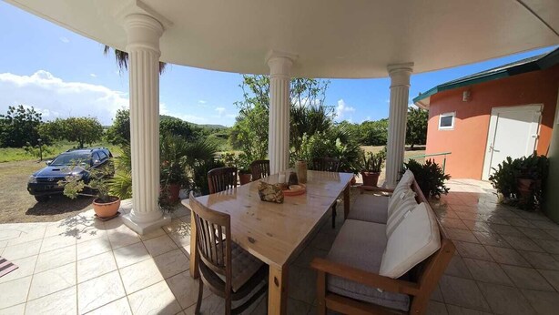 Un área de comedor al aire libre, techada, con una mesa y sillas de madera, rodeada de plantas en macetas y vistas a un coche y follaje.