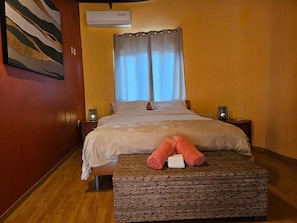 A hotel room with a bed, two bedside tables, a wall-mounted air conditioner, a window with curtains, and a painting on the wall.