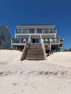 Una casa frente a la playa con balcón de dos pisos, escaleras de madera que conducen a la entrada y una playa arenosa al fondo.