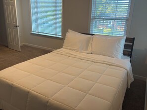 Una cama con ropa de cama blanca, dos ventanas con persianas, una puerta y suelo alfombrado.