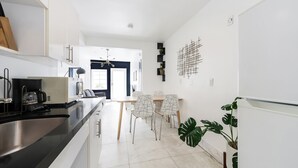 A modern kitchen with a black countertop, stainless steel sink, and a coffee maker. There are white cabinets, a dining area with a table and chairs, and a large plant. - Alton Apts by At Mine Hospitality (Miami Beach)