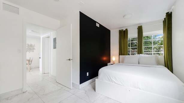 A modern hotel room with a large bed, a bedside table, a window with curtains, and a door leading to another room. - Alton Apts by At Mine Hospitality (Miami Beach)