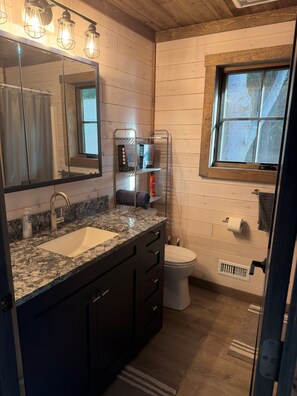 A bathroom with a granite countertop sink, a toilet, a window, and a shelf with toiletries. - New Build 2-bedroom 2-bath cabin on 2 acres -sleeps 8 near Silverlake Sand Dunes (Mears)