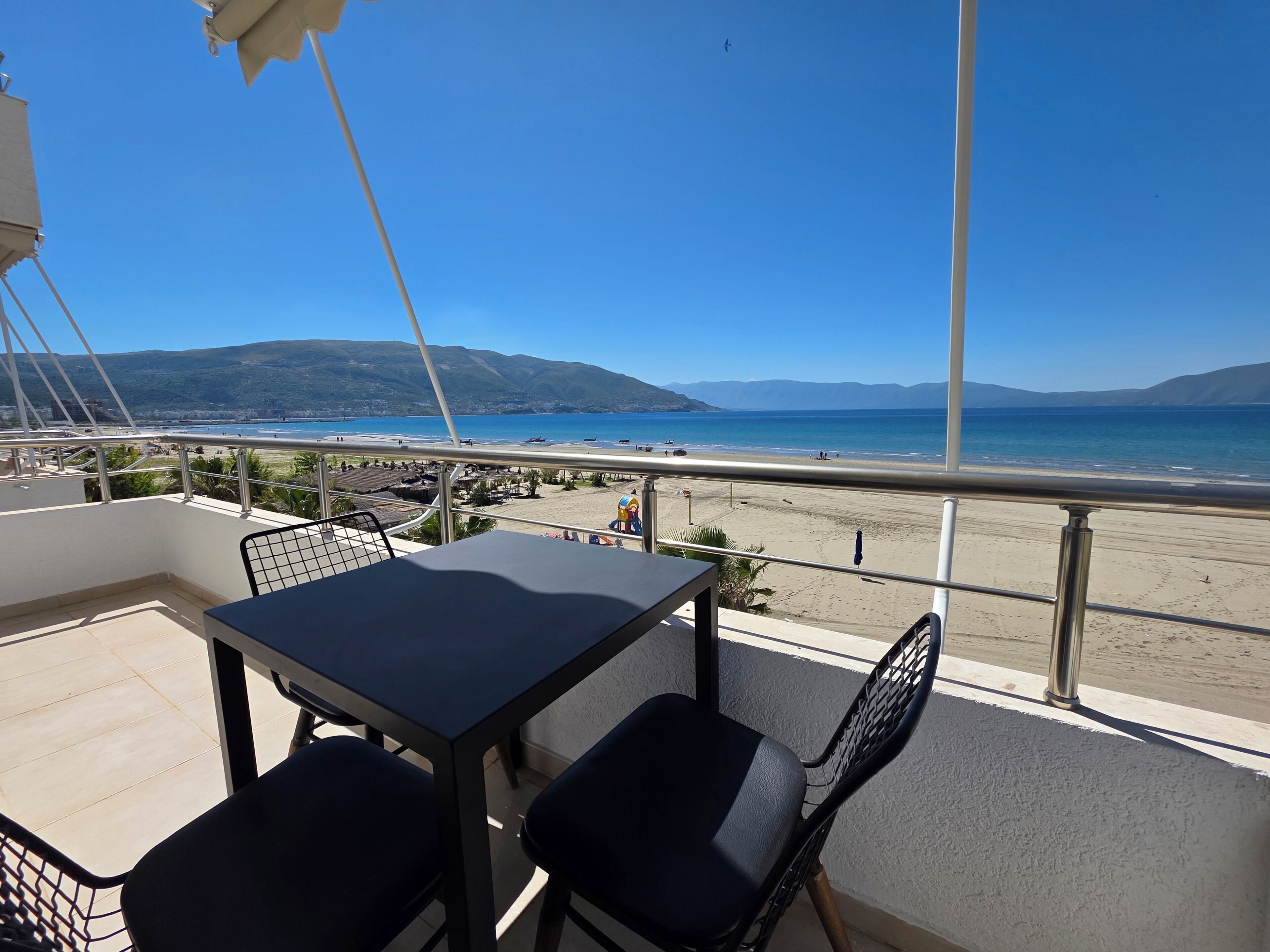 En balkon med et bord og stole, med udsigt over en strand og et klart hav med fjerne bjerge.