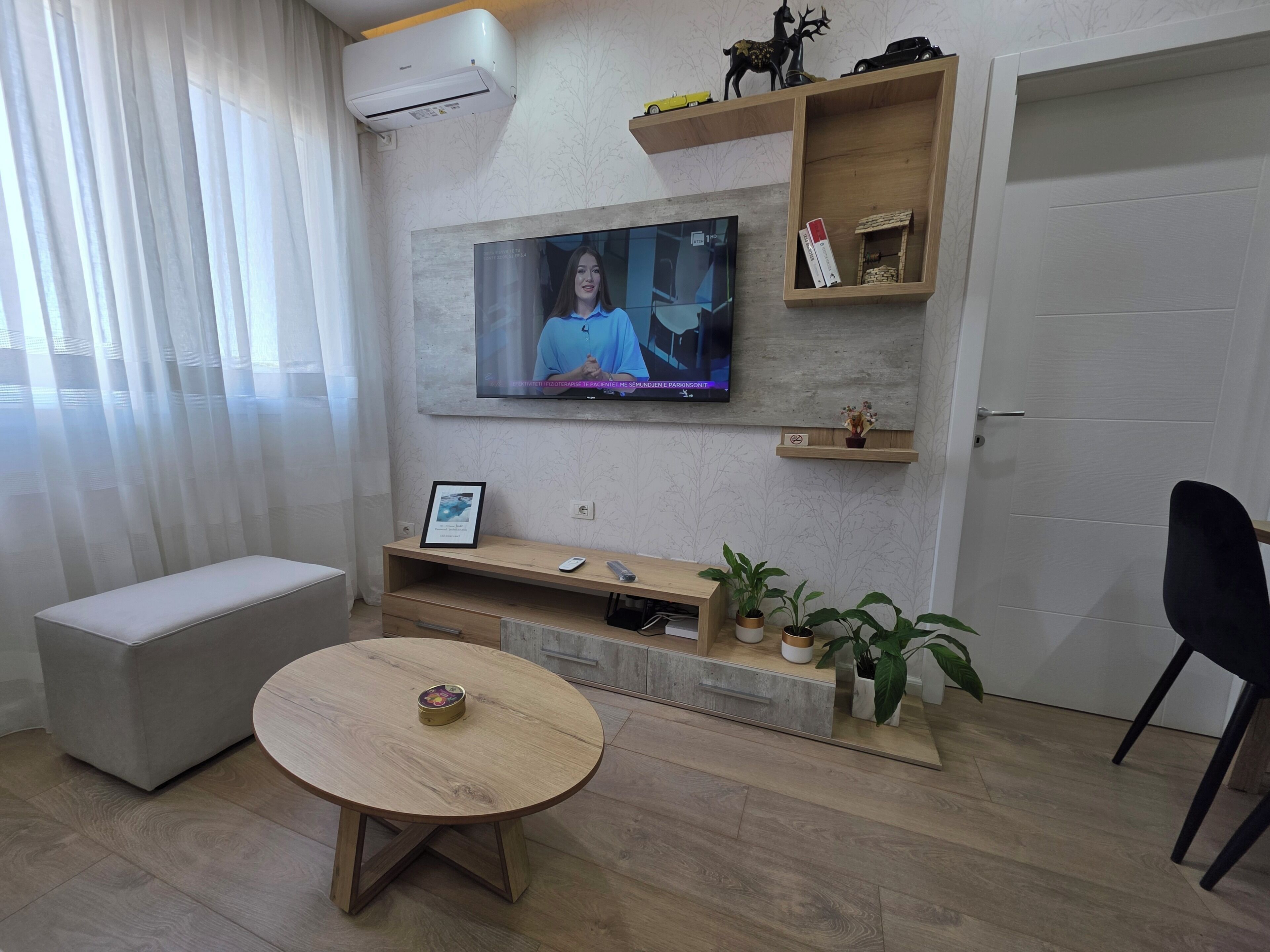 A modern living room with a flat-screen TV mounted on the wall, a round wooden table, a bench, and a black chair.