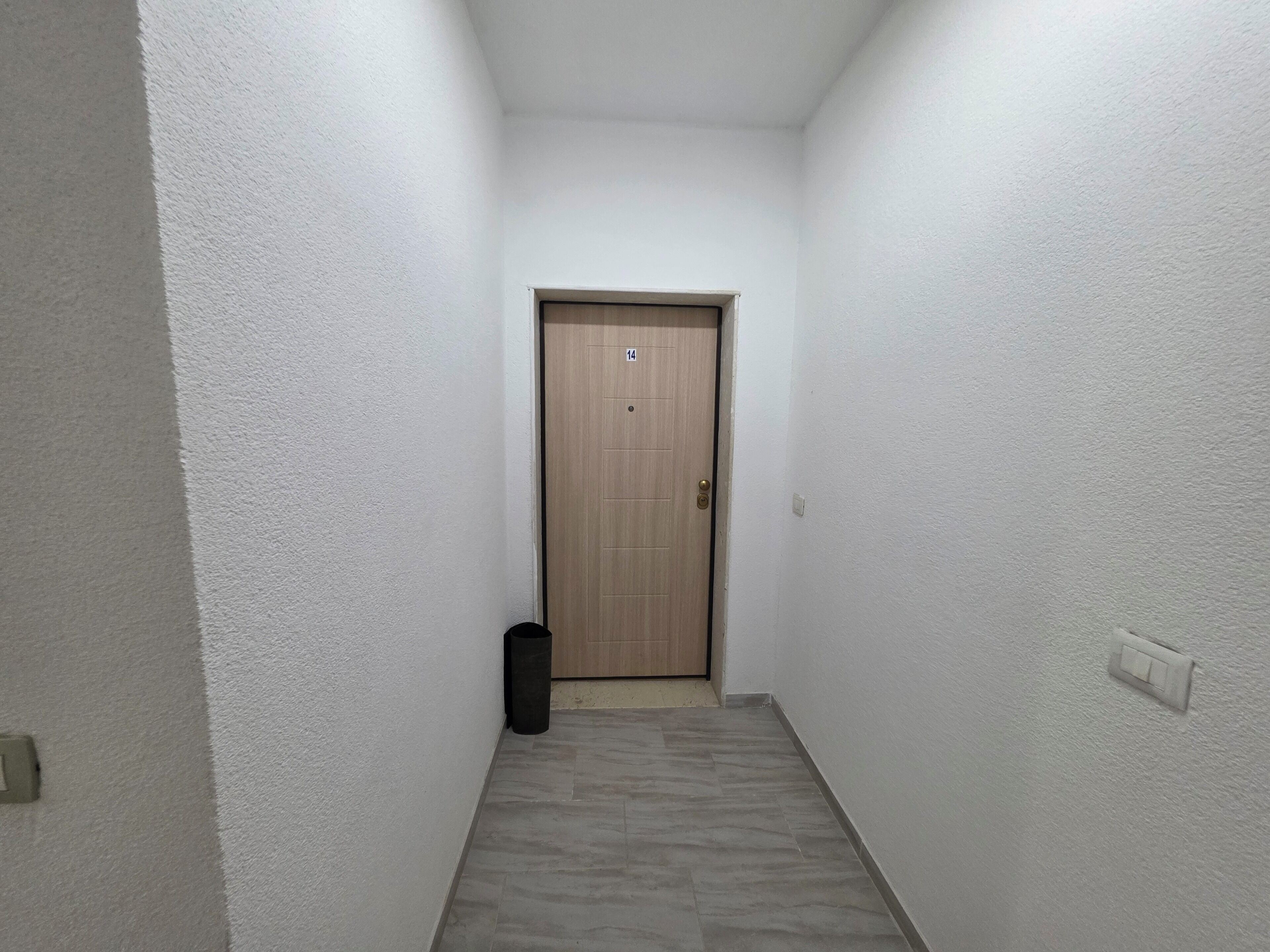 Passadís estret amb parets blanques, una porta de fusta, un cubell de brossa i un interruptor.