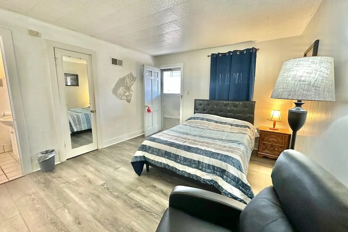 A bedroom with a bed, a nightstand, a lamp, and a door leading to another room.